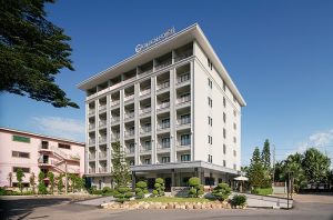 A modern six-story hotel building with balconies, labeled Golden Rover on the roof, surrounded by manicured shrubs and small trees under a clear blue sky.