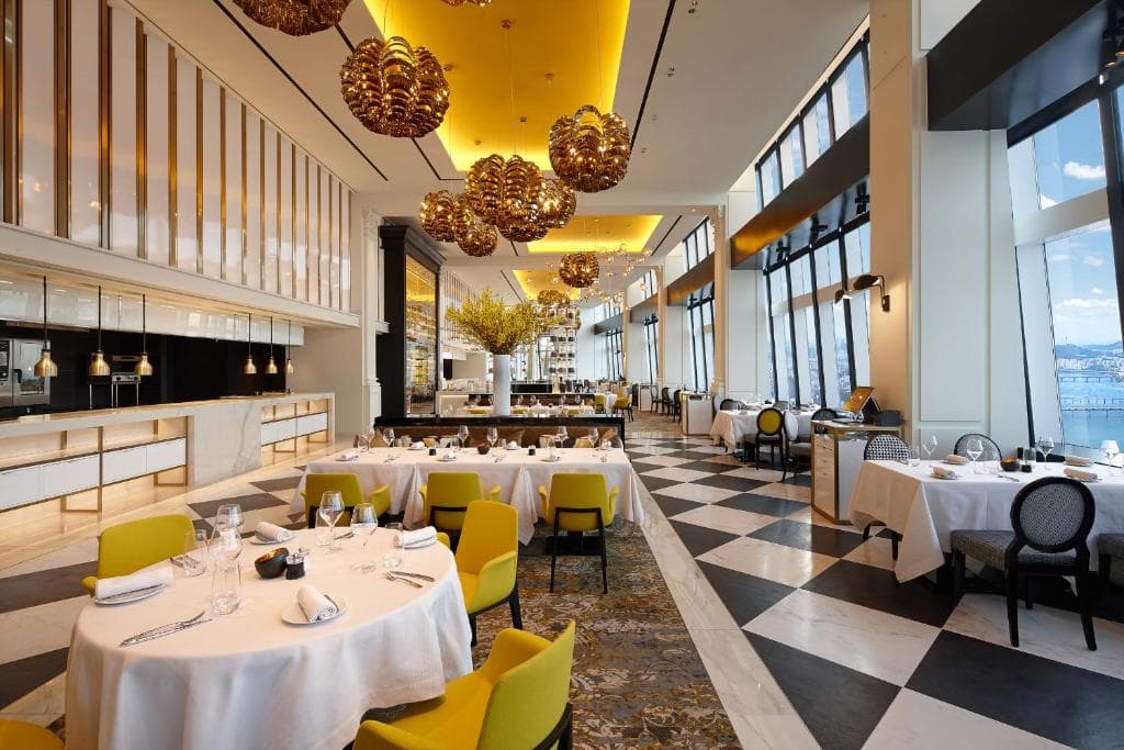 A modern, elegant restaurant with large windows, yellow chairs, round tables set with white tablecloths, and striking gold light fixtures hanging from a high ceiling. The space is bright and spacious with a checkered floor.