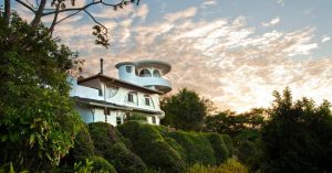 A unique white house with rounded and curved architectural features is nestled among lush green bushes and trees, with a dramatic, partly cloudy sky at sunset in the background.