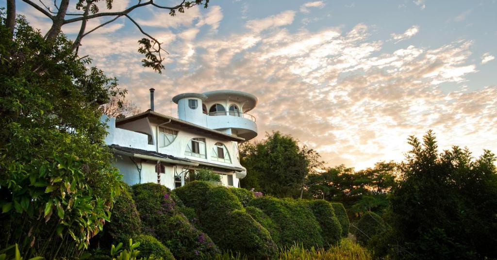 A unique white house with rounded and curved architectural features is nestled among lush green bushes and trees, with a dramatic, partly cloudy sky at sunset in the background.