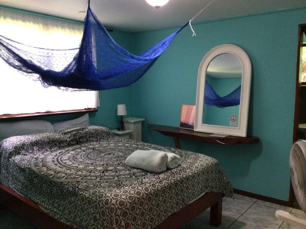 A cozy bedroom with teal walls, a bed covered in a patterned bedspread, a rolled towel on top, a blue mosquito net hanging above, a mirror on the wall, a small nightstand, and a window with sheer curtains.