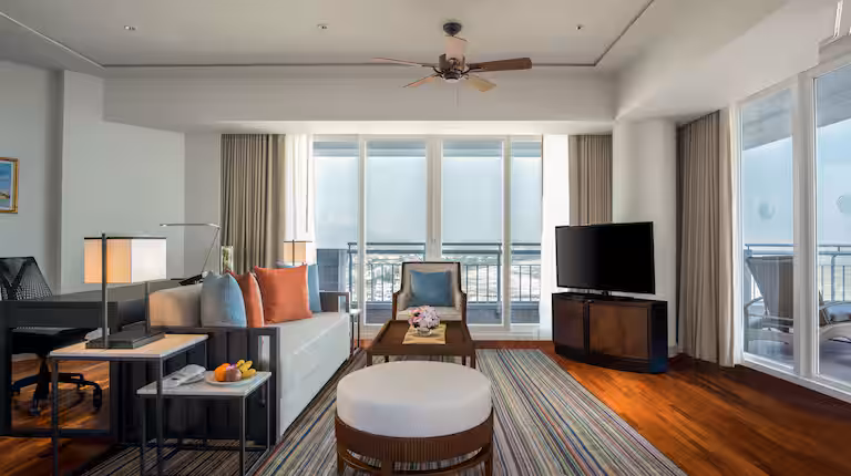 Modern hotel suite with hardwood floors, a striped rug, a sofa with colorful pillows, a coffee table, TV on a stand, desk, and large windows letting in natural light, leading to a balcony with outdoor seating.