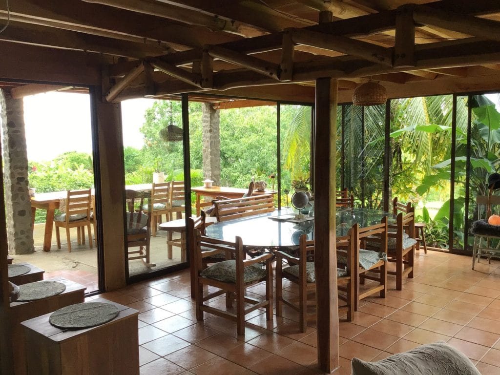 A spacious dining area with wooden chairs and a glass table sits next to large glass sliding doors that open to a lush, tropical garden and an outdoor patio with more wooden seating. Natural light fills the room.