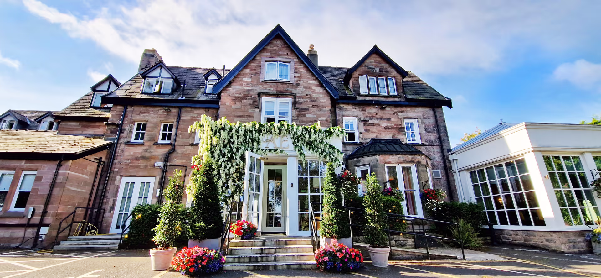 A charming stone building with large windows and a steep roof, adorned with hanging greenery above the entrance. Colorful flower pots decorate the steps leading to the main door on a sunny day.
