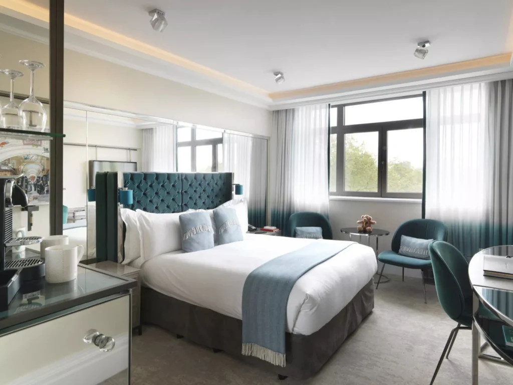 A modern hotel room with a double bed, teal headboard, blue throw and pillows, two armchairs by the window, a small table, a desk with a chair, and a coffee station on the left. Large windows let in natural light.