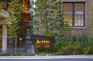 A wooden Ace Hotel sign stands in front of a tree and plants outside a brick building with large windows, near an entrance with warm lighting.