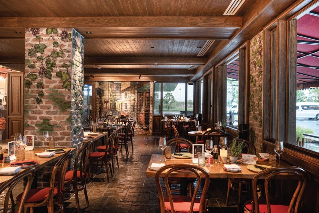 Cozy restaurant interior with wooden furniture, brick pillars decorated with grapevine murals, set tables, large windows letting in natural light, and a warm, inviting atmosphere.