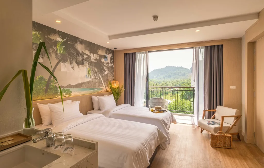 A bright hotel room with two neatly made twin beds, a tropical beach mural on the wall, a cozy chair, and a balcony door open to a scenic view of green hills and trees. Natural light fills the space.