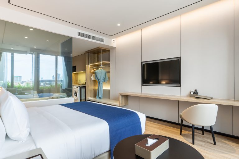 Modern hotel room with a large bed, blue blanket, glass wall, TV, long desk with chair, wardrobe with clothes, and a window showing city views. Room features minimalist decor and wooden flooring.