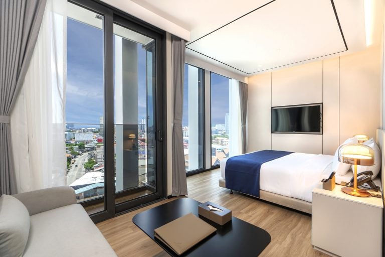 Modern hotel room with a large bed, sofa, coffee table, wall-mounted TV, and floor-to-ceiling windows offering a city view. Light-colored decor and natural light fill the space.