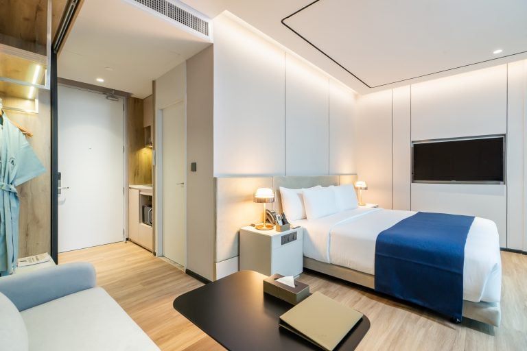 Modern hotel room with a double bed, white bedding, a blue throw, bedside tables with lamps, a wall-mounted TV, a small work desk, and a closet with robes. Light wood flooring and soft lighting create a cozy atmosphere.