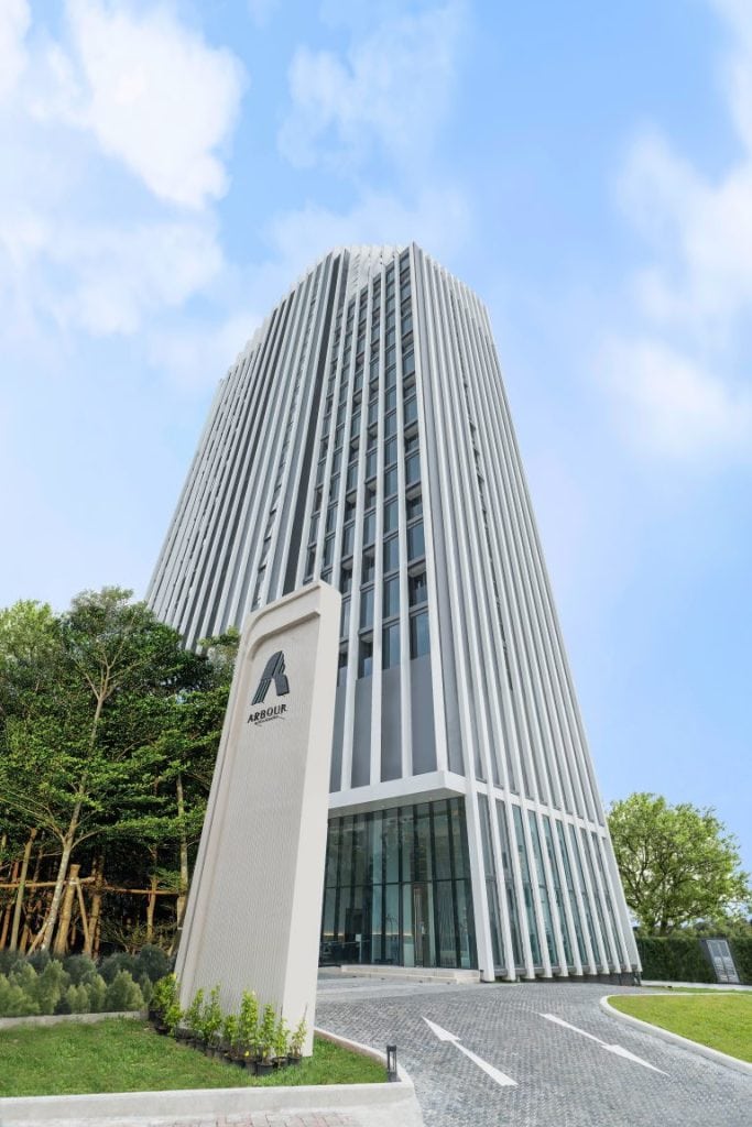 A modern glass and steel office building with vertical white fins, surrounded by green trees and blue sky. A sign with the logo and name AEON MALL stands in the foreground on a paved driveway.