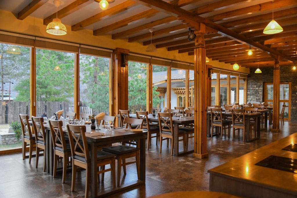 A modern restaurant dining area with wooden beams, large windows, wooden tables and chairs, set for guests, and views of greenery outside. Warm lighting creates a cozy and inviting atmosphere.