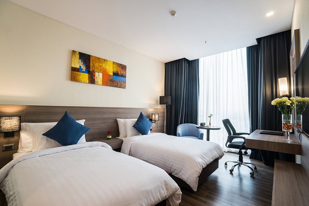 Modern hotel room with two single beds, blue accent pillows, a wooden desk with an office chair, large window with sheer curtains, wall art above the beds, and fresh flowers on the desk.