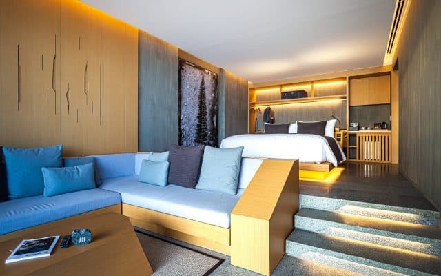 Modern hotel room with a sunken seating area featuring a light blue sectional sofa, a wooden coffee table, steps leading up to a bed with white linens, and warm ambient lighting throughout.