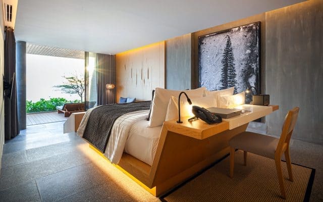 Modern hotel room with a large bed, wooden desk, chair, and soft lighting. A large framed tree artwork hangs on the wall. Floor-to-ceiling windows reveal greenery and an outdoor patio area.