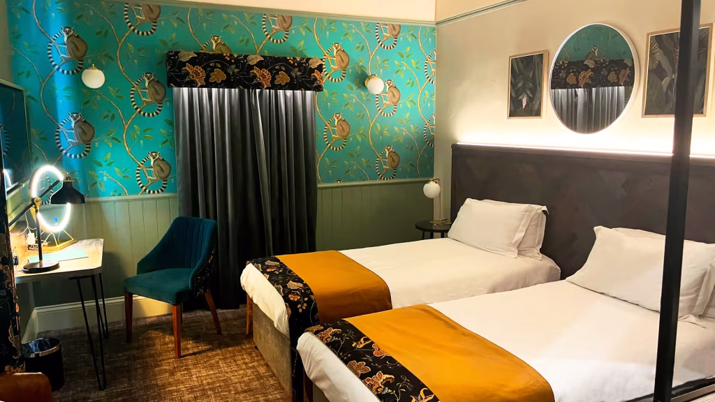 A stylish hotel room with two single beds, white bedding, and gold accents. The room has a teal chair, a desk with a mirror, patterned teal wallpaper, and decorative wall art above the beds.