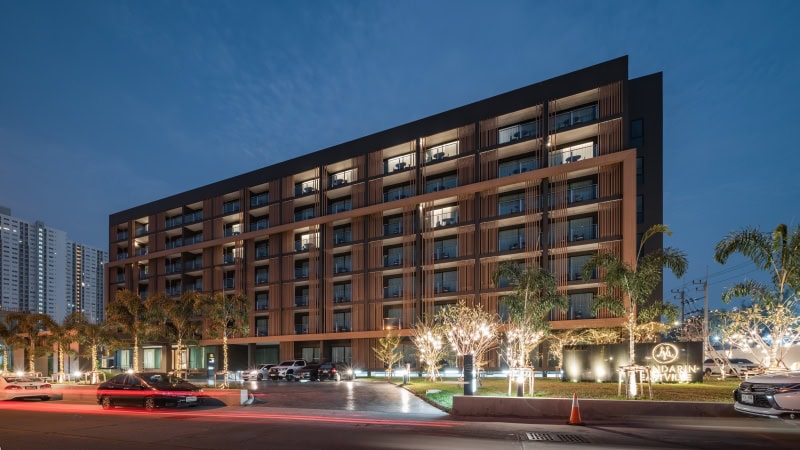 A modern, multi-story apartment building with illuminated balconies and ground-level lights, surrounded by palm trees and parked cars, photographed at dusk.