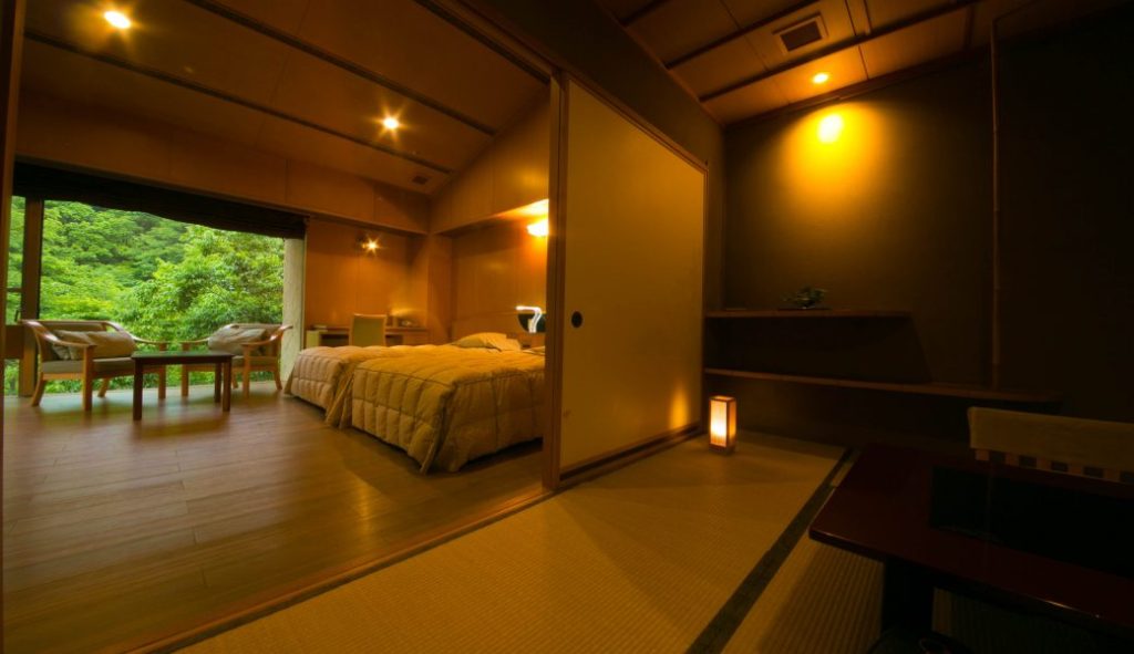 A warmly lit Japanese-style hotel room with tatami flooring, sliding doors, a low table, and a bedroom area featuring two beds and large windows overlooking green forest scenery.