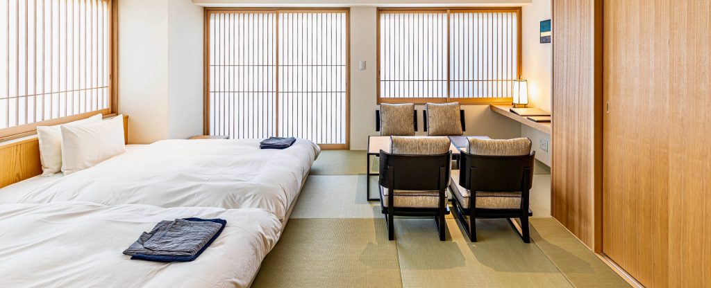 A bright, minimalist Japanese-style room with two beds, a low table with four chairs, tatami mats, and large shoji screens letting in natural light. A lamp and decorative items are on a side counter.