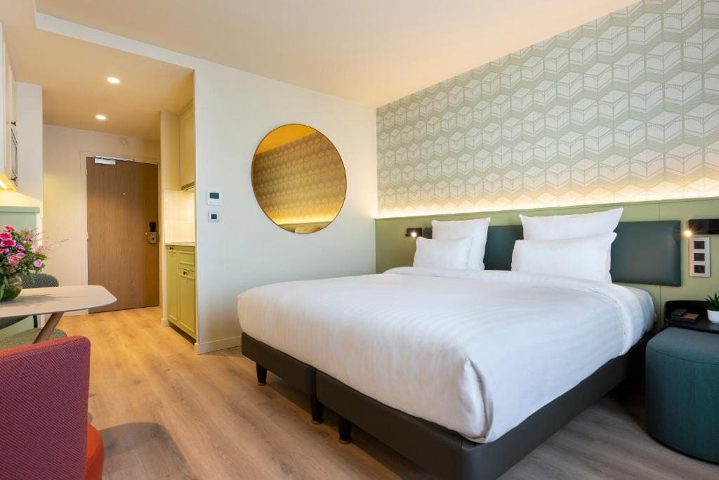 A modern hotel room with a large double bed, white bedding, geometric patterned accent wall, round mirror, small desk with a red chair, flowers in a vase, and light wood flooring near an open entryway.