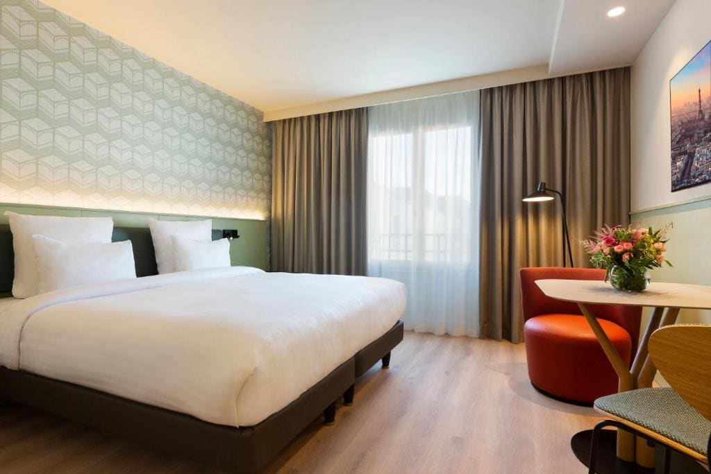Modern hotel room with a large bed, geometric wallpaper, a small round table with two chairs, a bouquet of flowers, a tall window with sheer and blackout curtains, and a wall-mounted photo.