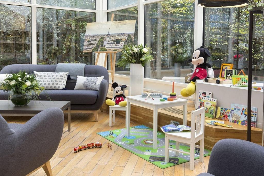 A bright, cozy playroom with large windows, gray sofas, a small white table and chair, toys, books, two Mickey Mouse plushies, a play mat with roads, and a toy train on a wooden floor.