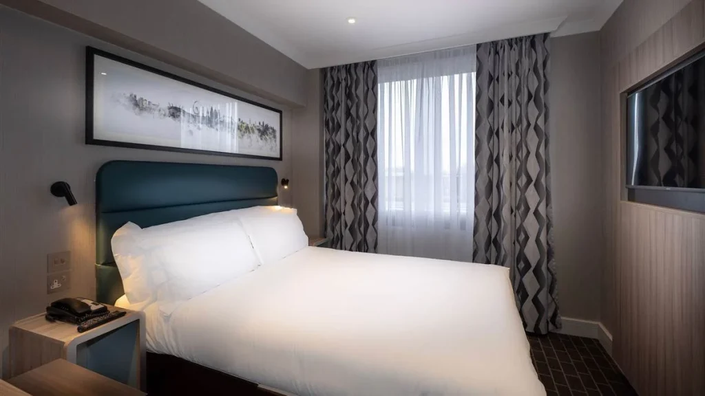A modern hotel room with a double bed, white bedding, wall-mounted lamps, a phone on the bedside table, patterned curtains, a large window, and a wall-mounted TV.
