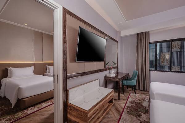 Modern hotel room with two double beds, a flat-screen TV mounted on the wall, a desk with a chair, a window with curtains, and an adjacent bedroom visible through a sliding door. Neutral and wood tones dominate the decor.