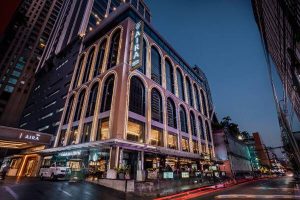 A modern multi-story hotel with large arched windows and bright lighting, situated on a busy city street at dusk. The hotel sign reads AIRA and vehicles are visible in front of the building.