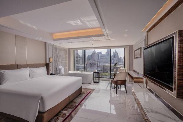 Modern hotel room with a large bed, a bathtub near floor-to-ceiling windows, a desk, a TV, and a city skyline view outside. The room has marble floors and elegant, neutral décor.