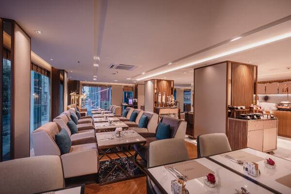 Modern, elegant restaurant interior with neatly arranged tables and cushioned chairs, soft lighting, wood accents, and decorative pillows. Large windows line one side, and a buffet area is visible in the background.