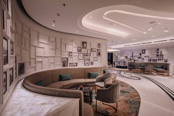 Modern hotel lounge with curved sofa, armchairs, and a marble table. The walls are decorated with a grid of framed photos, and the space is softly lit with recessed ceiling lights. The decor is elegant and contemporary.