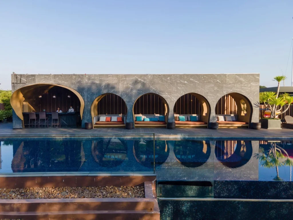 A modern building with large arched openings and colorful seating overlooks a clear blue swimming pool, reflecting the arches and sky, with a bar area visible on the left.