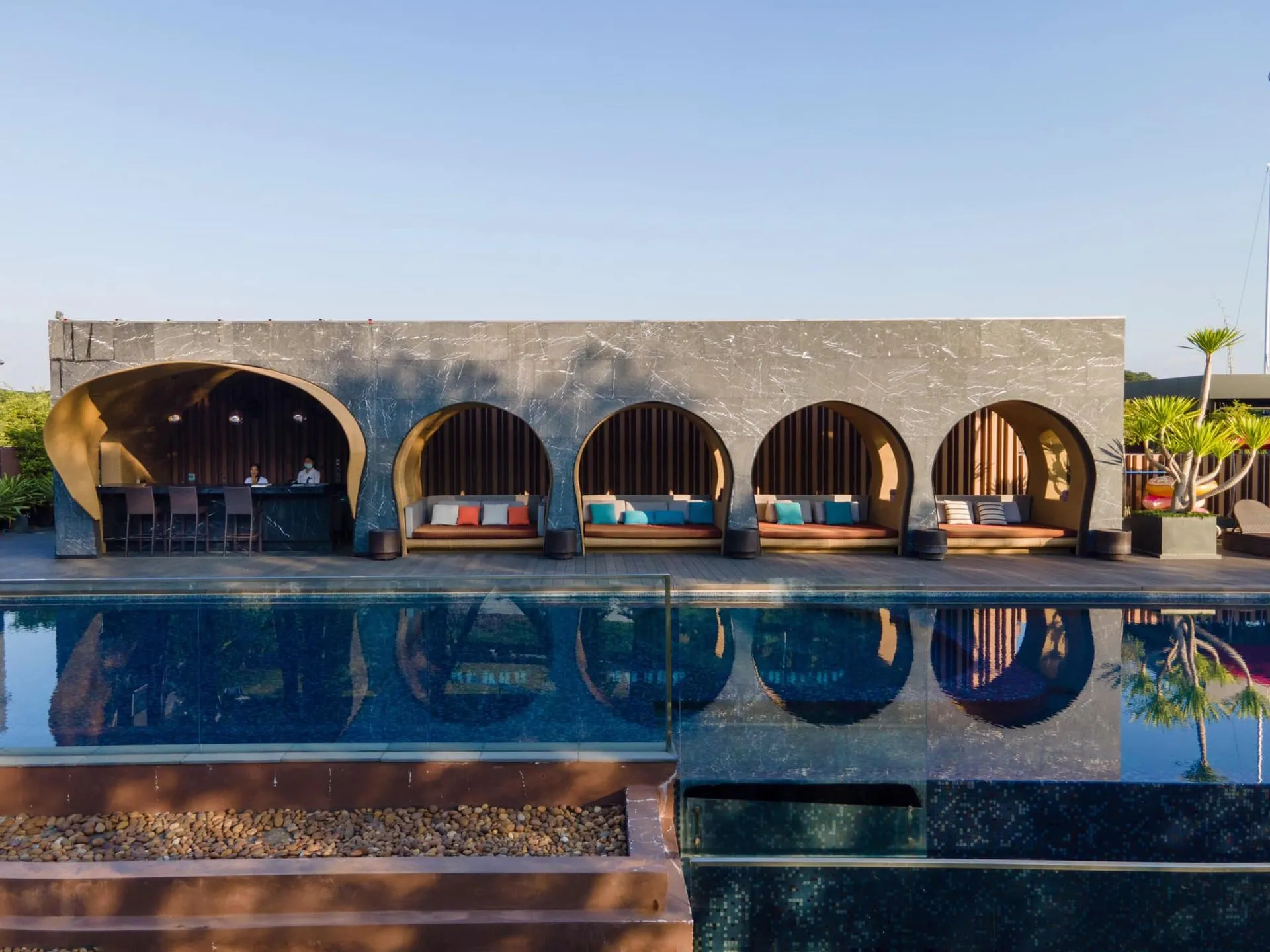 A modern building with large arched openings and colorful seating overlooks a clear blue swimming pool, reflecting the arches and sky, with a bar area visible on the left.