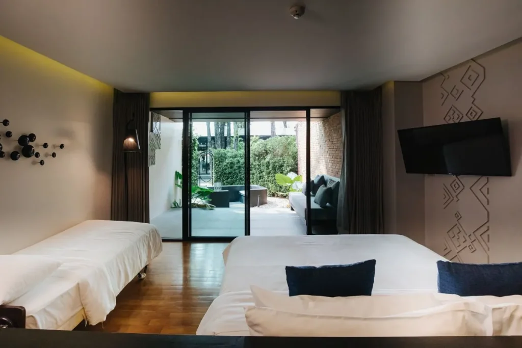 Modern hotel room with two beds, a wall-mounted TV, and decorative wall art. Glass doors open to a patio with outdoor seating and greenery, letting in natural light. The room has wood flooring and a cozy, minimalist design.