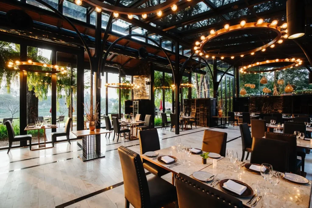 A modern, elegant restaurant with glass walls and ceiling, large circular chandeliers, neatly set tables with black chairs, and views of lush greenery outside at sunset.
