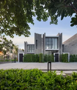 A modern two-story house with tall, vertical concrete panels, large glass windows, and greenery along the front. Trees and shrubs frame the scene, and a paved area is visible in the foreground.