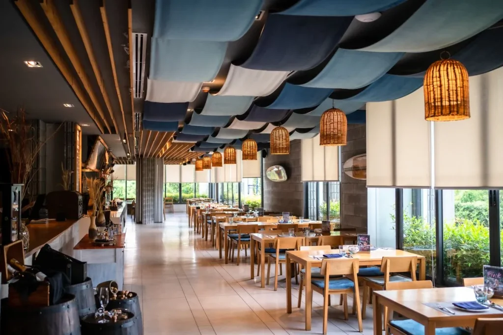 A modern restaurant with wooden tables and chairs, rattan pendant lights, and a ceiling decorated with blue and white fabric waves. Large windows let in natural light and offer views of greenery outside.