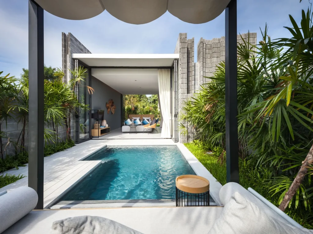 Modern outdoor pool area with lush greenery, lounge seating, and a covered patio leading to a glass-walled indoor living space with contemporary decor and natural light.