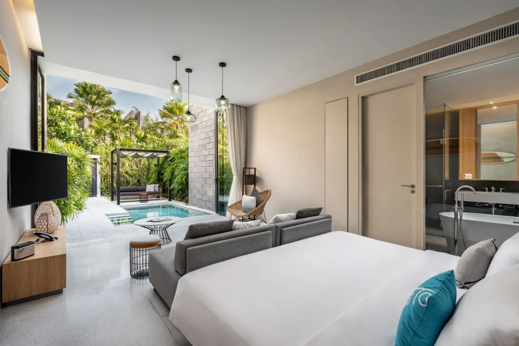 A modern hotel room with a large bed, sofa, TV, and glass wall opens to a private patio with a pool surrounded by lush greenery and outdoor seating, creating a luxurious and tranquil atmosphere.