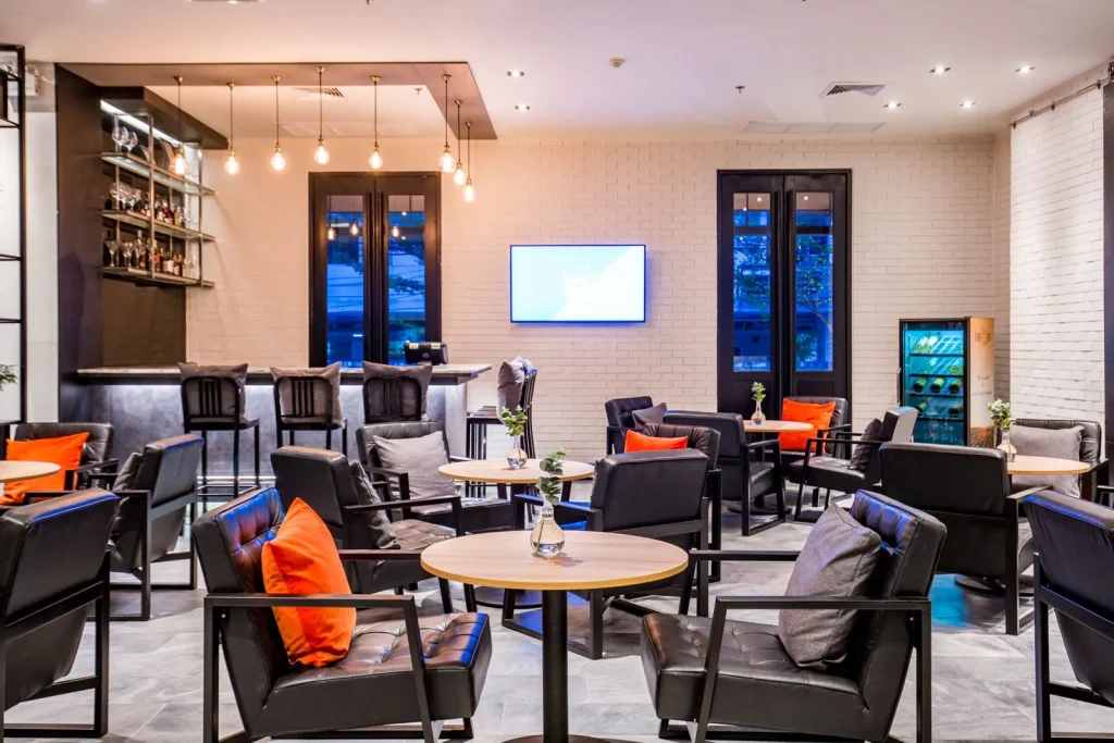 Modern café interior with black chairs, orange and gray cushions, round wooden tables, pendant lighting, a wall-mounted TV, and a bar area with shelves of bottles. Large windows and a vending machine are visible.