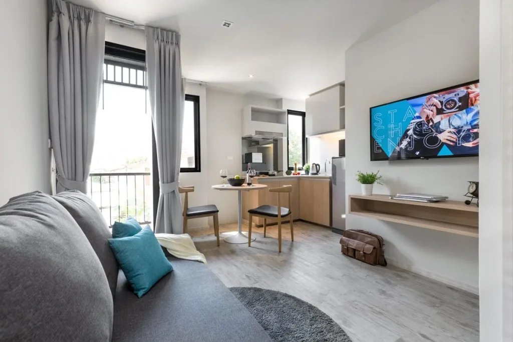 Modern, bright studio apartment with a gray sofa, blue and teal pillows, round dining table with two chairs, wall-mounted TV, kitchen area, large windows with gray curtains, and light wood flooring.
