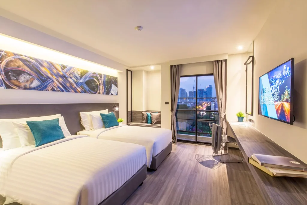 A modern hotel room with two neatly made twin beds, teal accent pillows, a wall-mounted TV, a work desk, and a large window overlooking a cityscape at dusk. The room is brightly lit and stylishly decorated.