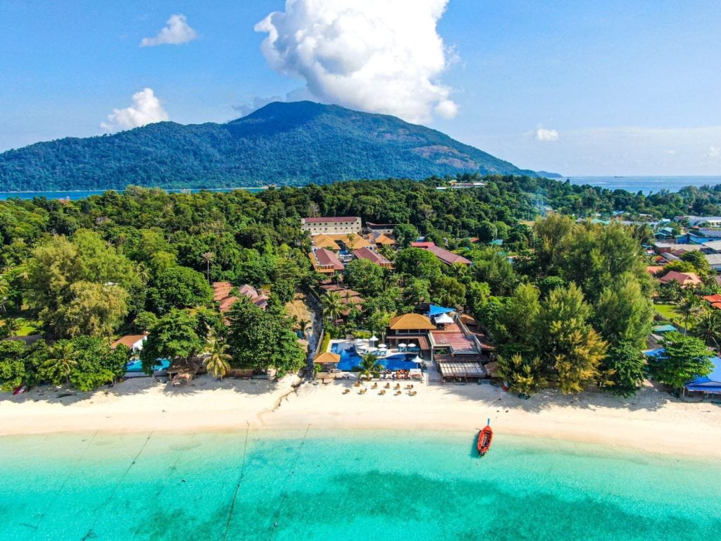 Aerial view of a tropical beach with turquoise water, white sand, a small red boat near shore, lush green trees, and buildings, with a forested mountain and cloudy blue sky in the background.