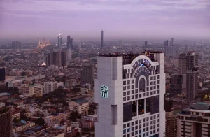A tall, modern high-rise with a rooftop bar overlooks a sprawling cityscape at dusk, with buildings and lights stretching into the hazy distance under a purple sky.