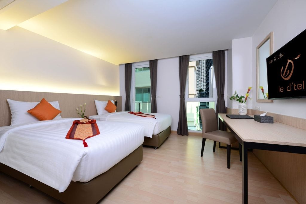 A modern hotel room with two neatly made twin beds, each with white linens and orange accent pillows, a desk with a chair, a TV mounted on the wall, and a large window with curtains letting in natural light.