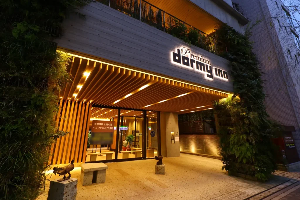 The entrance of a brightly lit Premium Dormy Inn hotel at night, with wooden slats, glass doors, greenery on the walls, and two animal statues flanking a bench near the entrance.