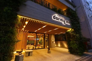 The entrance of a brightly lit Premium Dormy Inn hotel at night, with wooden slats, glass doors, greenery on the walls, and two animal statues flanking a bench near the entrance.