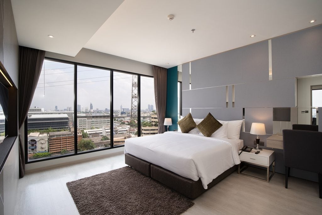 Modern hotel room with a large bed, bedside tables with lamps, a small desk and chair, large floor-to-ceiling windows with city views, and neutral-colored decor with brown and gray accents.